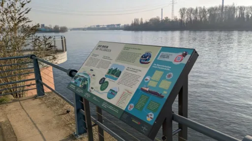 Informationstafel „Der Rhein bei Wiesbaden" am Rheinufer mit Blick auf den Fluss und das gegenüberliegende Industrieufer